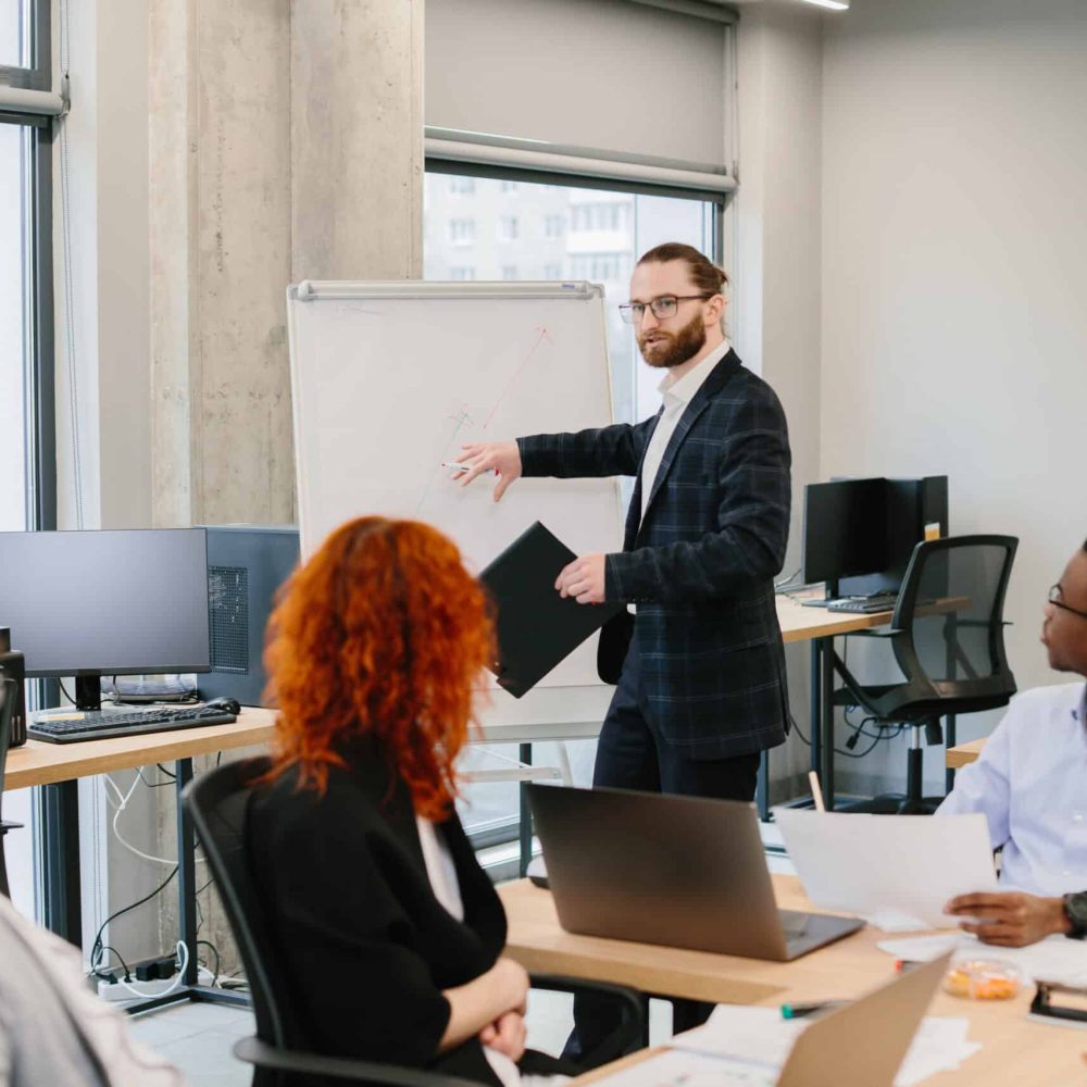 Businessman leads a presentation using a whiteboard, engaging with a diverse team of professionals in a contemporary office setting
